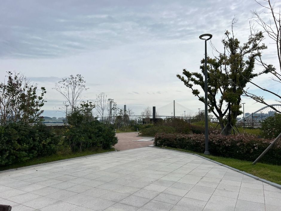 Paved plaza with shrubs, young trees and a lamp post in a seaside park, a cable-stayed bridge on the cloudy horizon