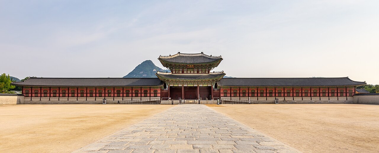 Heungnyemun Gate at Gyeongbokgung Palace with long red-pillared wings, a stone path across an empty sandy plaza, mountain behind