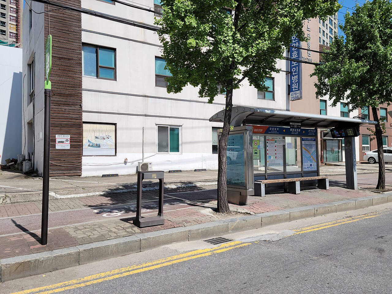 Empty roadside bus shelter with a bench, route maps and an arrival display, beside trees and a white building on a sunny day
