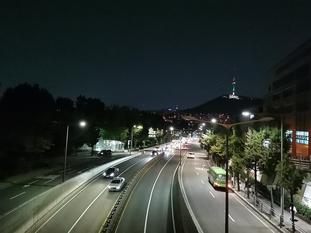 Multi-lane city road at night with streetlights, light traffic, a green bus and a lit tower on a hill in the distance