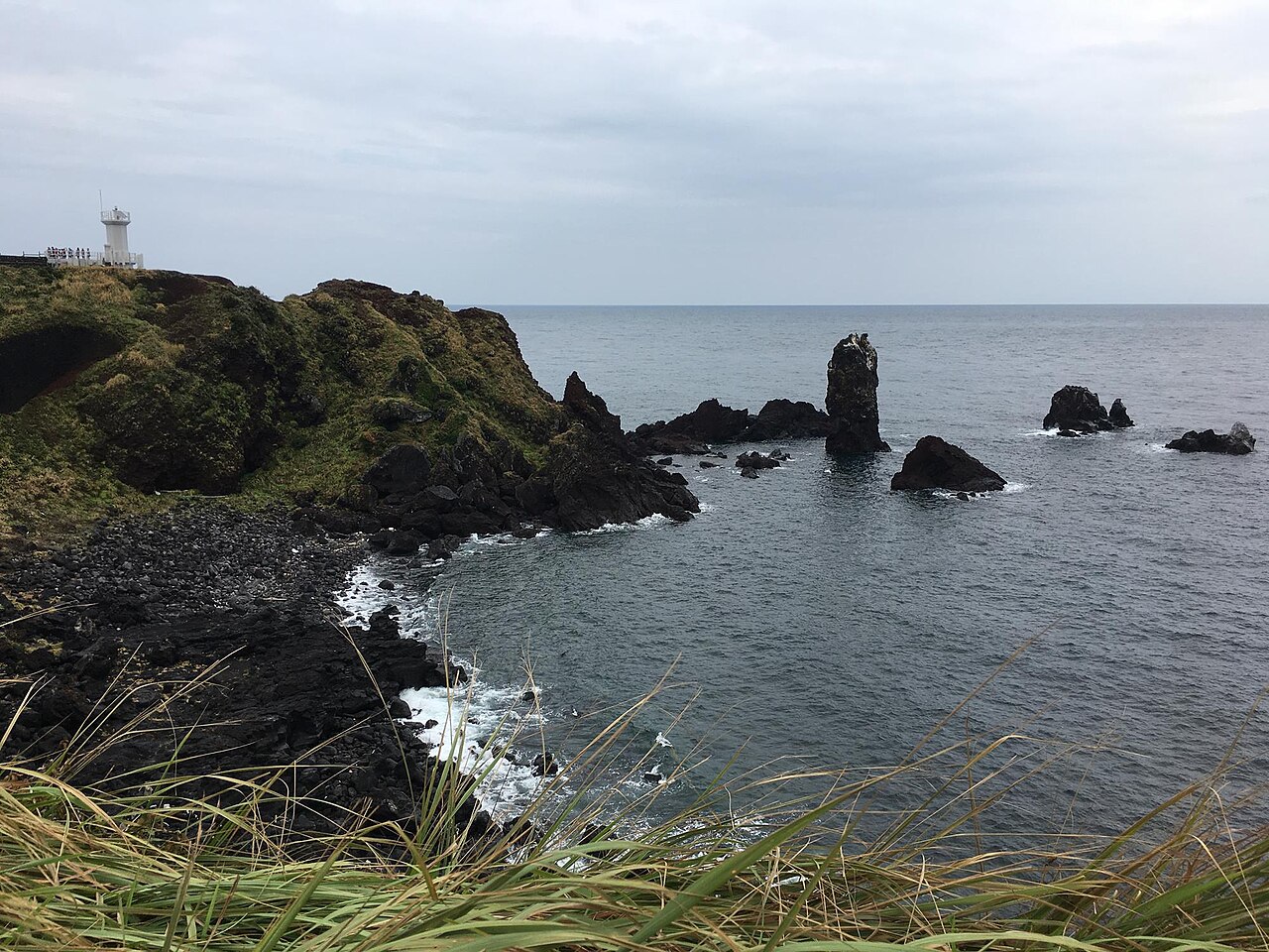 Dark volcanic cliffs and sea stacks on a grey sea under an overcast sky, with a small white lighthouse on the headland