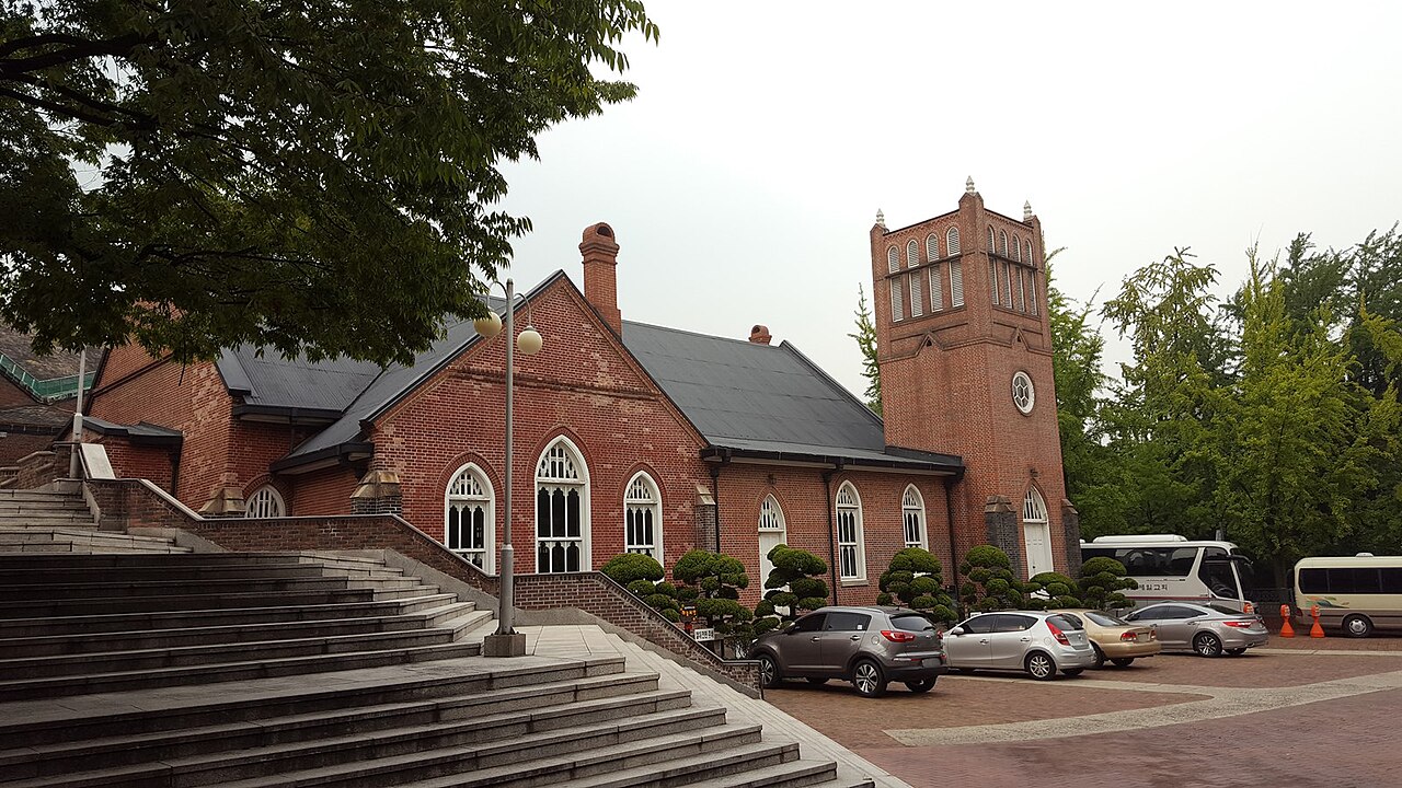 Red-brick church with arched windows and a square bell tower beside wide stone steps, cars parked in front, trees around
