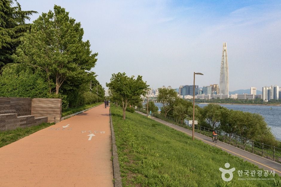 Tree-lined riverside path and grassy bank along the Han River at Ttukseom, with Lotte World Tower on the skyline