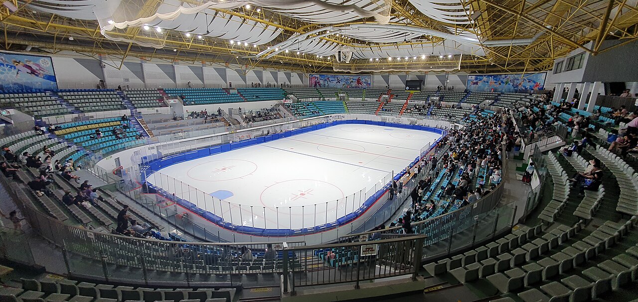 Indoor ice rink with a white rink, blue boards, tiered teal seats and a distant crowd of spectators under a yellow steel roof